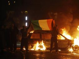 Nacionalistas extienden la bandera de Irlanda frente a unos autos que están quemándose, en Belfast. AP  /