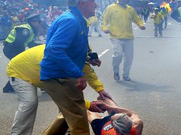 Un corredor es auxiliado por agentes de seguridad tras el primer estallido. Al fondo se aprecia la segunda detonación. AP /