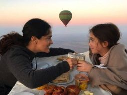 En la imagen se ve a ambas de perfil ante una mesa, con un globo aerostático, el mar y el amanecer al fondo. INSTAGRAM / salmahayek