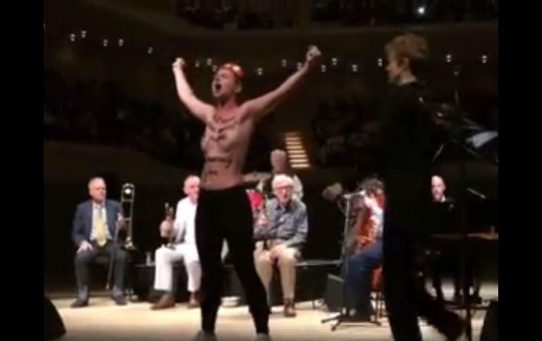 Una mujer con el torso desnudo y frases escritas en negro sobre la piel sube al escenario y grita con los brazos en alto. FACEBOOK / FEMEN