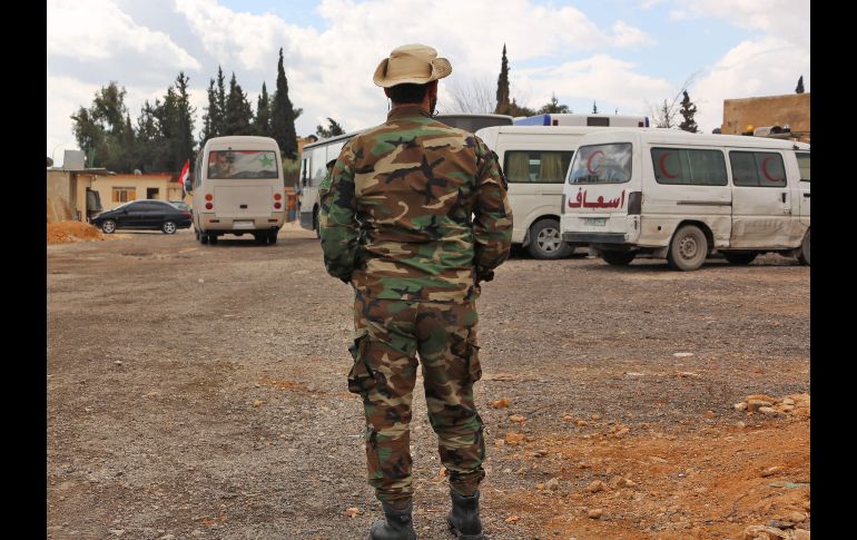 Un elemento de las fuerzas del régimen sirio se para junto a camiones y ambulancias que se utilizarán para el traslado de civiles.