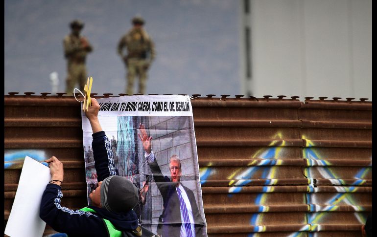 Un manifestante coloca un cartel en una valla en la frontera. Del lado estadounidense, elementos de seguridad vigilan junto a un prototipo.