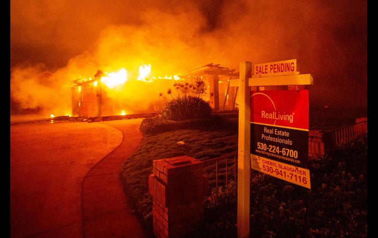 Un letrero de venta de propiedad se ve frente a una casa en llamas.