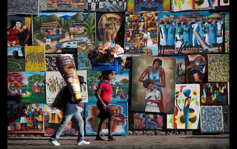 Un vendedor de remedios y una vendedora de caramelos caminan frente a pinturas típicas haitianas en Puerto Príncipe, Haití. EFE/O. Barría