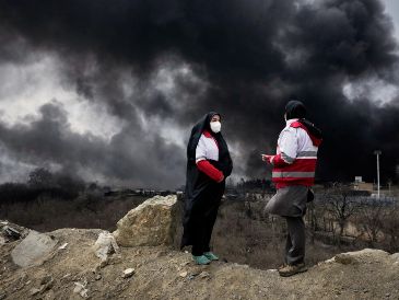 Dos mujeres de la Sociedad de la Media Luna Roja iraní permanecen de pie mientras una densa columna de humo proveniente de un ataque estadounidense-israelí contra una instalación de almacenamiento de petróleo en Teherán, Irán. AP/V. Salemi