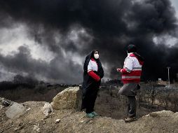 Dos mujeres de la Sociedad de la Media Luna Roja iraní permanecen de pie mientras una densa columna de humo proveniente de un ataque estadounidense-israelí contra una instalación de almacenamiento de petróleo en Teherán, Irán. AP/V. Salemi