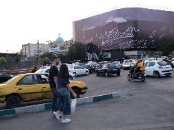 Una mujer y un hombre cruzan a unos metros de un enorme mural que muestra a Donald Trump en un ataúd, acompañada de una frase en persa que dice: 