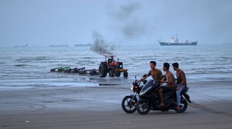 Jóvenes a bordo de motocicletas a lo largo de la orilla mientras un tractor saca equipo del agua y al fondo se pueden ver buques mercantes aparentemente anclados en el estrecho de Ormuz. AP/R. Poudat