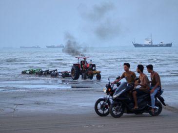 Jóvenes a bordo de motocicletas a lo largo de la orilla mientras un tractor saca equipo del agua y al fondo se pueden ver buques mercantes aparentemente anclados en el estrecho de Ormuz. AP/R. Poudat