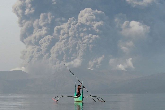 TOPSHOT - A youth living at the foot of Taal volcano rides an outrigger canoe while the volcano spews ash as seen from Tanauan town in Batangas province, south of Manila, on January 13, 2020. The Philippines was on alert January 13 for the 